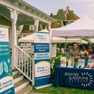A community event booth for the Allergy & Asthma Network is set up outdoors with banners, brochures, and people gathered near a tent and a house. Directional signs point to the 2026 event. Several people are talking at the table.
